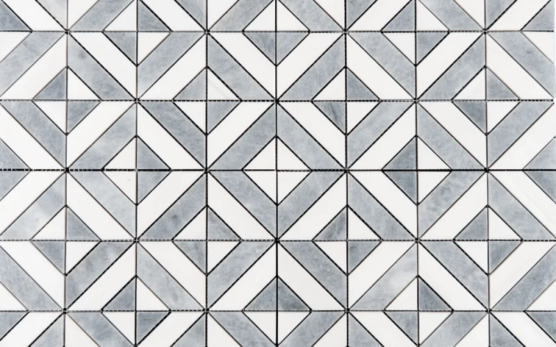 A geometric mosaic tile design in white and gray tones. The tile features a diamond and square pattern formed by small, intricately cut triangular and rectangular pieces.